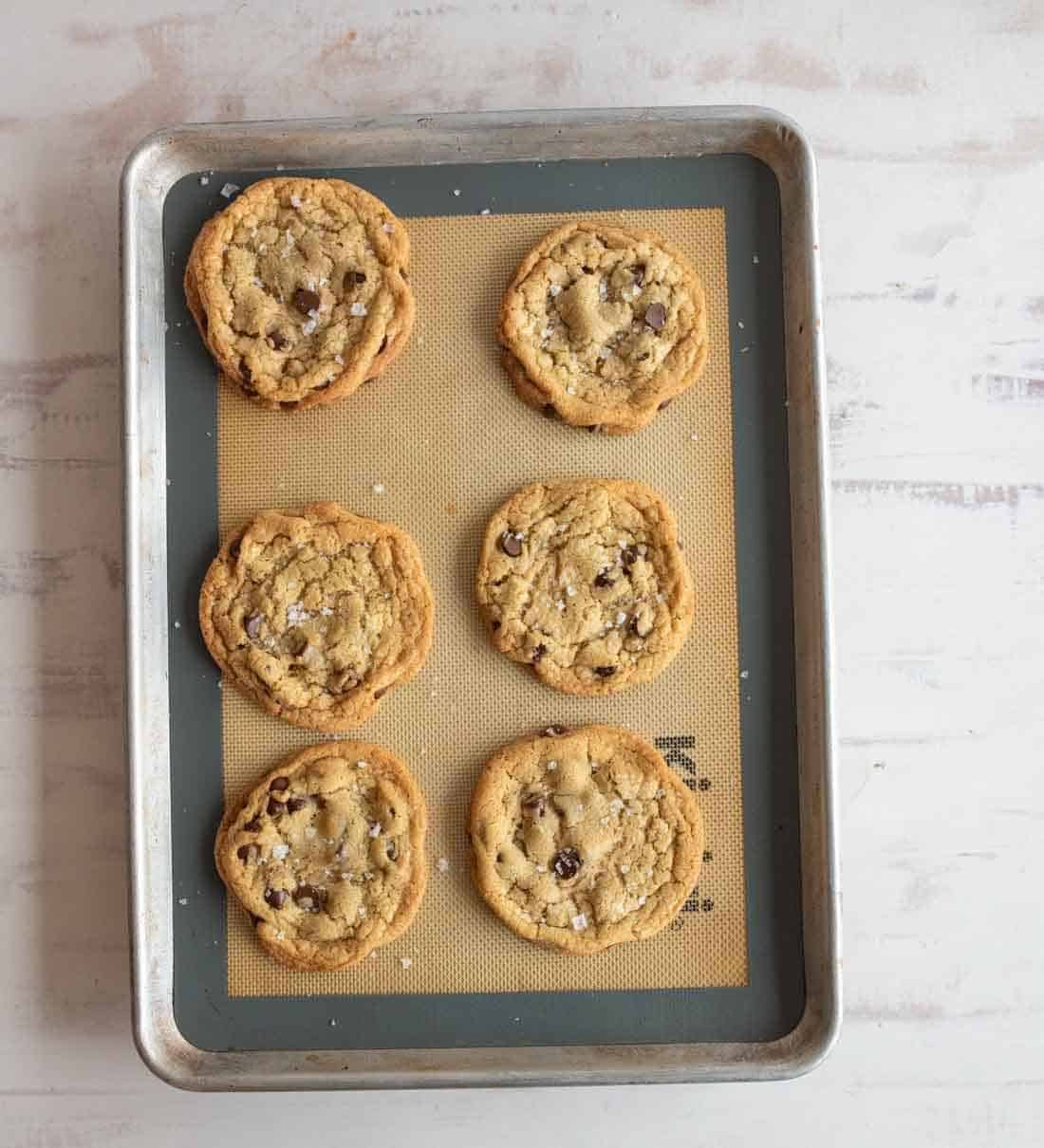 A baking sheet with six freshly baked chocolate chip cookies on a silicone mat, placed on a light, textured surface. The cookies are golden brown and sprinkled with a touch of salt.