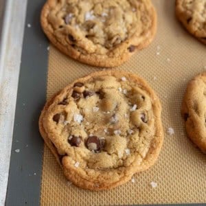 Close-up of freshly baked chocolate chip cookies on a baking sheet. The cookies are golden brown, with visible chocolate chips, and sprinkled with sea salt, resting on a beige silicone baking mat.