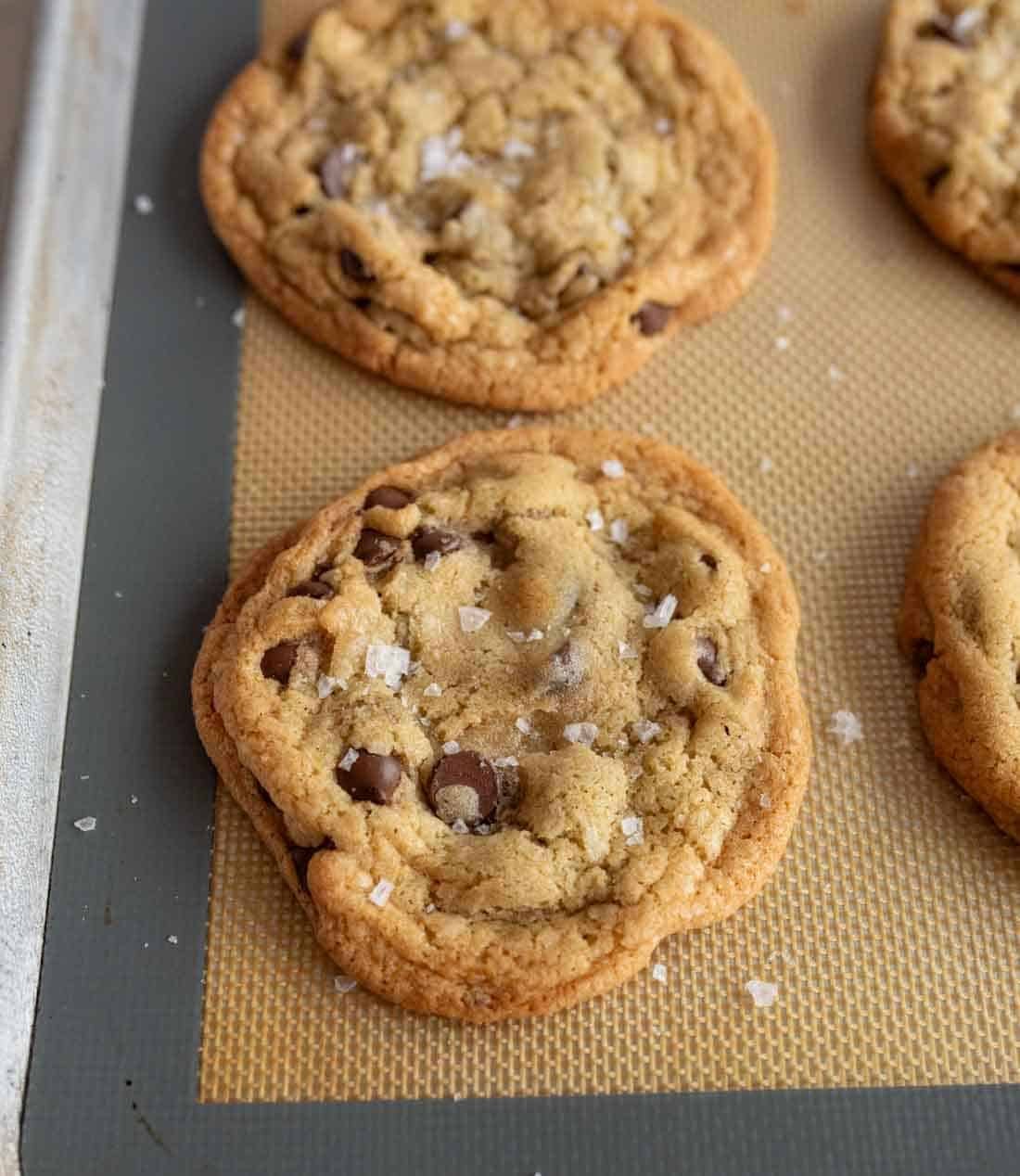 Close-up of freshly baked chocolate chip cookies on a baking sheet. The cookies are golden brown, with visible chocolate chips, and sprinkled with sea salt, resting on a beige silicone baking mat.