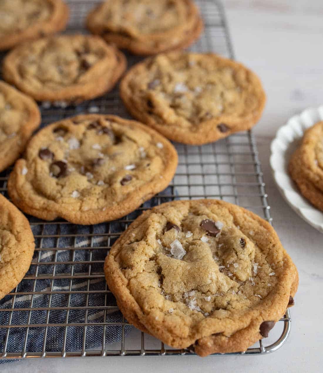 Chocolate chip cookies with a sprinkle of sea salt cooling on a wire rack, placed on a white surface. Some cookies are on a white plate nearby. The cookies appear freshly baked and golden brown.