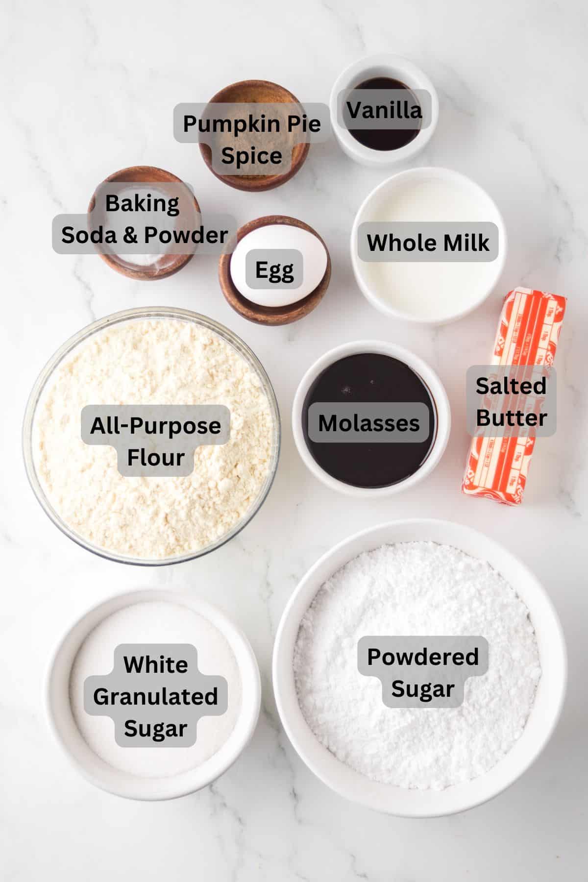 Baking ingredients are arranged on a white surface: labeled bowls of all-purpose flour, powdered sugar, granulated sugar, egg, vanilla, whole milk, molasses, baking soda and powder, pumpkin pie spice, and a stick of salted butter.