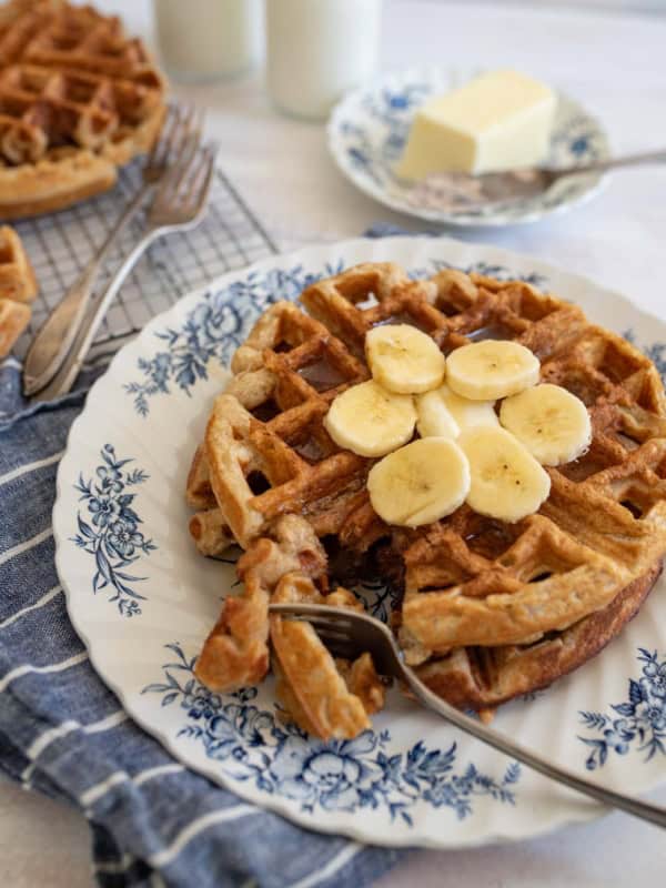 A plate with banana oatmeal waffles topped with banana slices, placed on a blue floral-patterned plate. A fork rests on the waffle. A pat of butter is on a plate in the background, with a blue cloth and other waffles nearby.