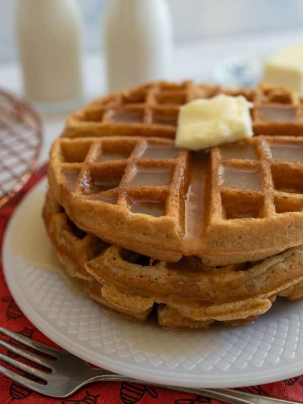 A stack of cinnamon waffles topped with a pat of butter sits on a white plate with syrup drizzled over them. In the background, there are two milk bottles, a block of butter, a fork, and a red napkin.