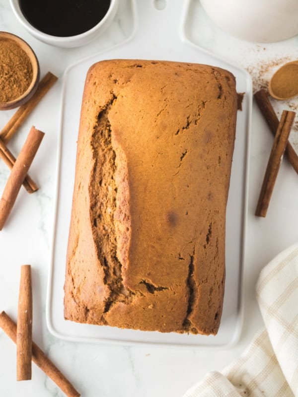 Freshly baked gingerbread loaf rests on a white cutting board, surrounded by cinnamon sticks and a small bowl of cocoa powder. A cup of dark liquid and a white cloth are nearby, set on a white marble surface.