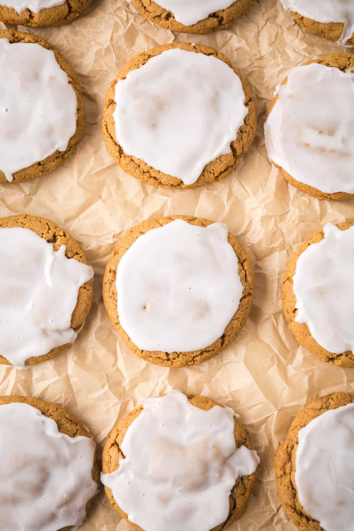 Iced cookies arranged in rows on crinkled parchment paper. The cookies have a light brown color with white icing covering the tops. The icing is slightly uneven, giving a homemade look.