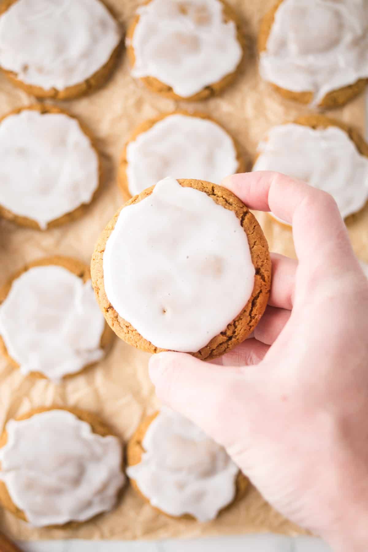 A hand holding a brown cookie topped with white icing. In the background, several similar cookies are arranged on parchment paper.