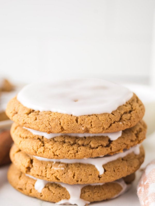 A stack of five iced gingerbread cookies on a white surface. Each cookie is topped with a thin layer of white icing, and the edges are slightly uneven, adding a homemade touch. The background is softly blurred.