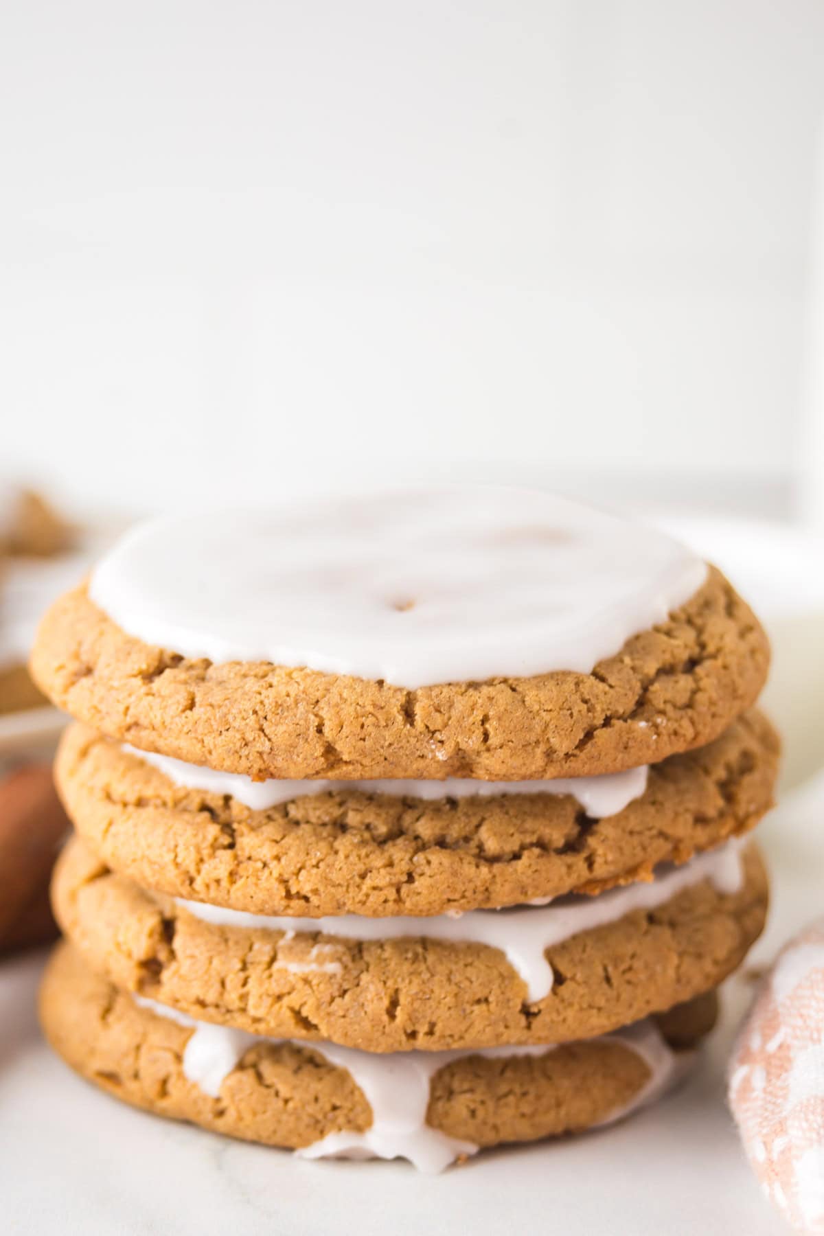 A stack of five iced gingerbread cookies on a white surface. Each cookie is topped with a thin layer of white icing, and the edges are slightly uneven, adding a homemade touch. The background is softly blurred.