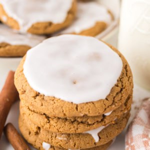 A stack of gingerbread cookies topped with white icing, next to cinnamon sticks. A partially visible plate of more cookies is in the background, along with a bottle of milk and a patterned cloth.