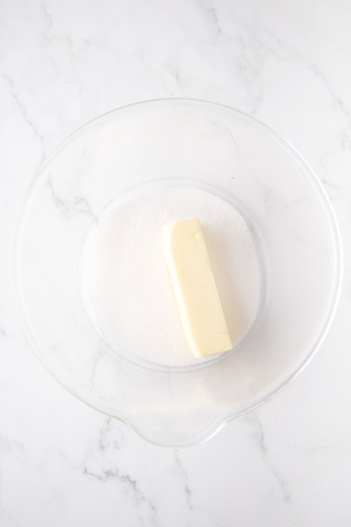 A stick of butter and a pile of white sugar sit in a transparent glass mixing bowl. The bowl is placed on a white marble surface.