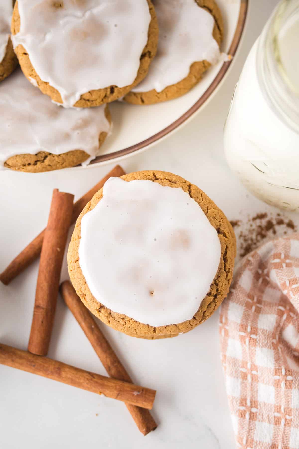 Frosted cookies on a plate and a single frosted cookie on a table alongside cinnamon sticks. A patterned cloth napkin and a jar with a white liquid, likely milk, are also visible.