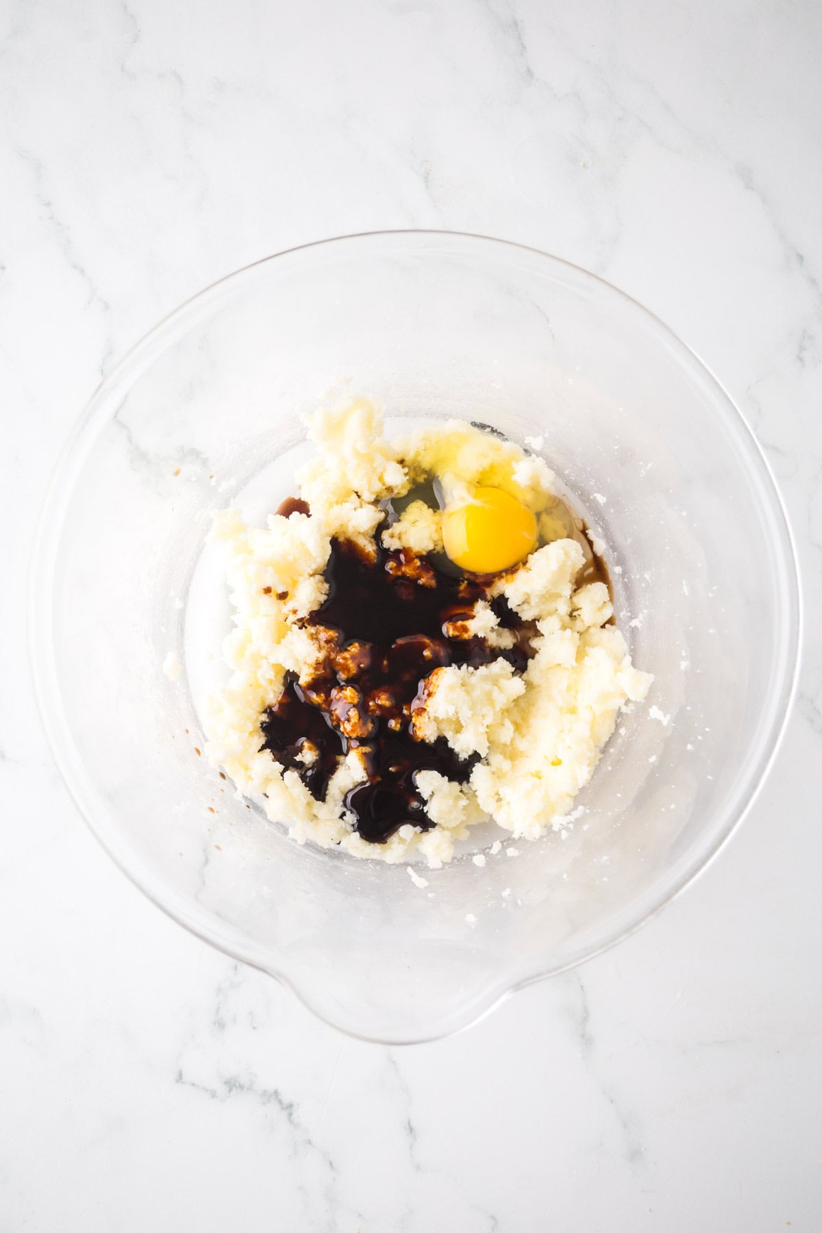 A glass bowl containing a mixture of butter and sugar with an egg cracked in. Vanilla extract has been added, creating pools of brown liquid on the mixture. The background is a white marble surface.