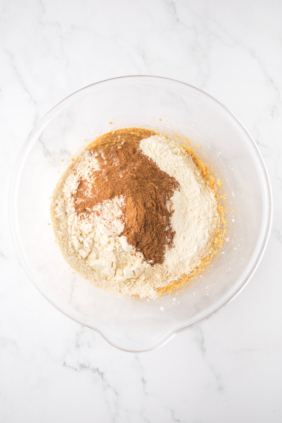 A clear mixing bowl containing flour, cocoa powder, and sugar on a marble countertop. The ingredients are unmixed, with the cocoa and flour creating a contrast in color.