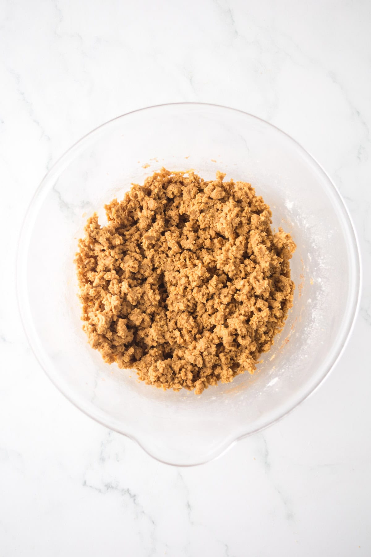 A glass bowl on a marble surface filled with a crumbly brown mixture, resembling cookie dough or crumble topping.