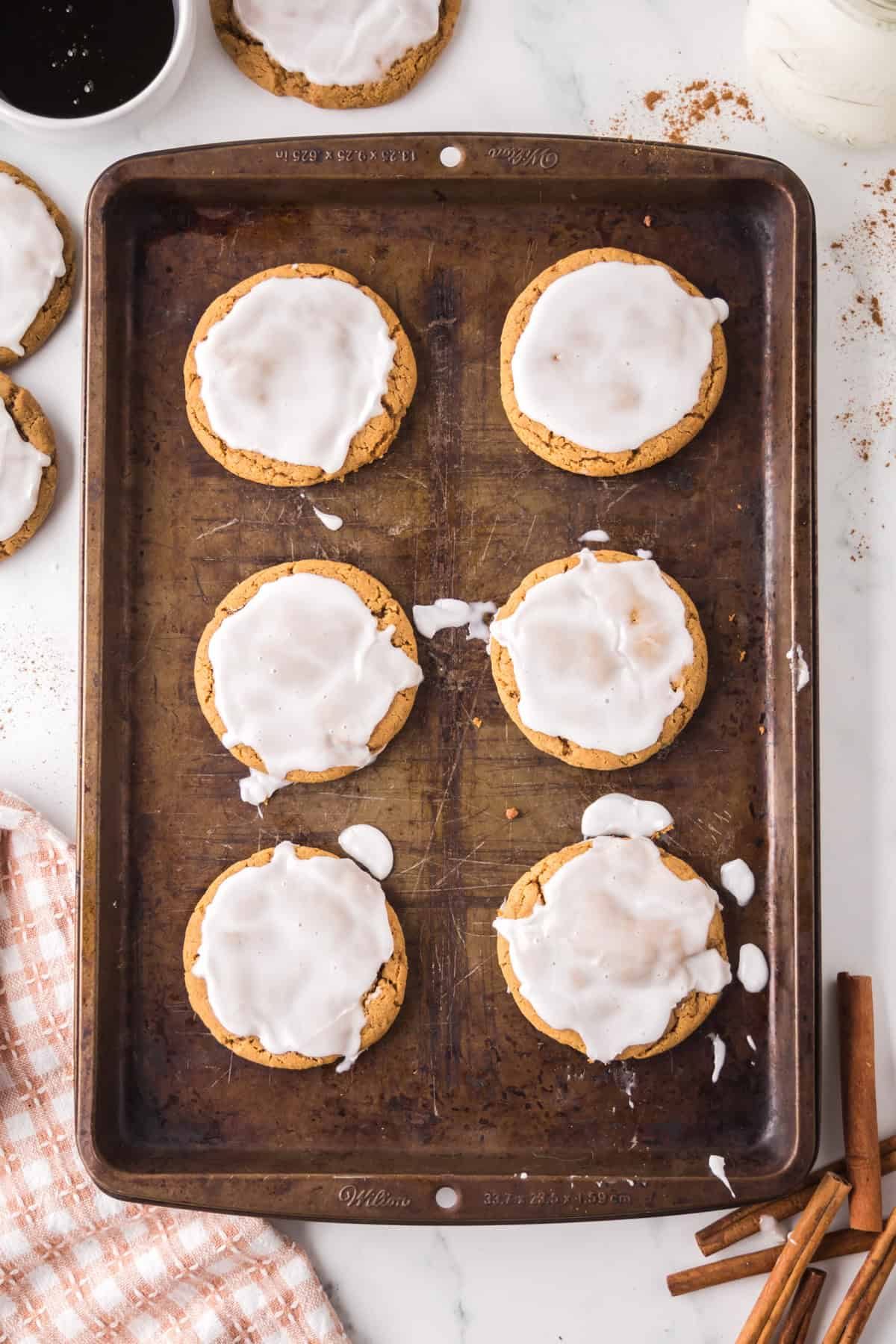 A baking tray holds six round cookies topped with white icing. Some icing has dripped onto the tray. Two additional cookies, a checkered cloth, cinnamon sticks, and a black bowl with sauce are beside the tray.